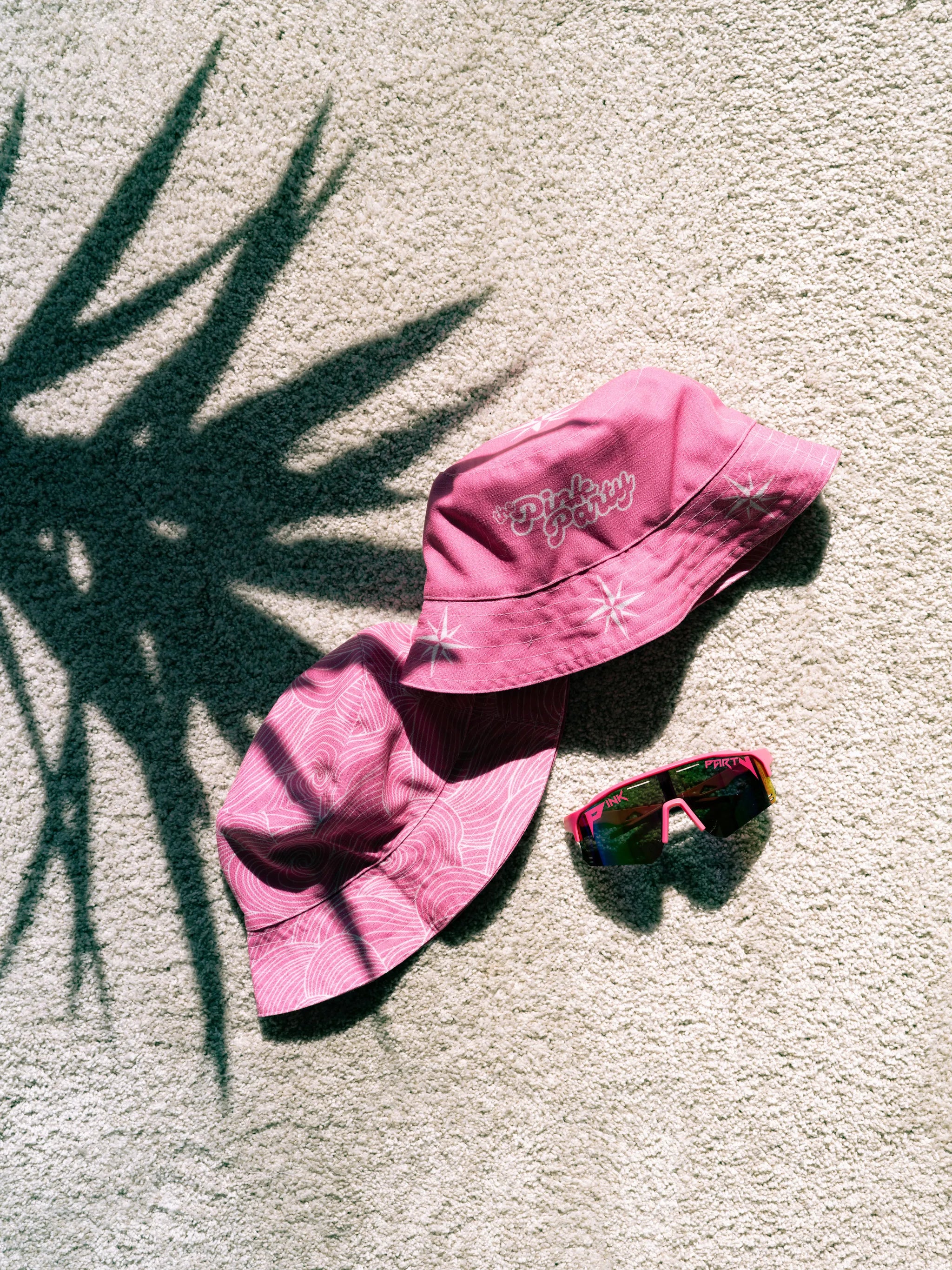 Two pink bucket hats and a pair of sunglasses on a textured surface with a shadow from a plant.
