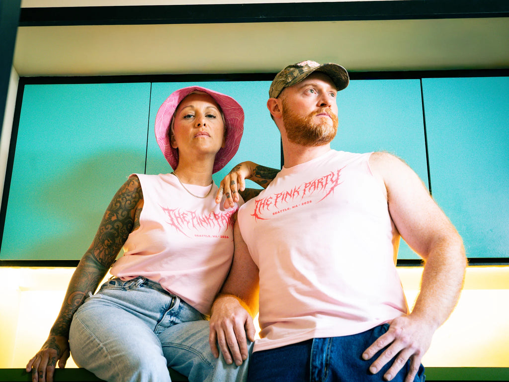 Two people wearing matching pink tank tops with text, sitting against a teal wall.