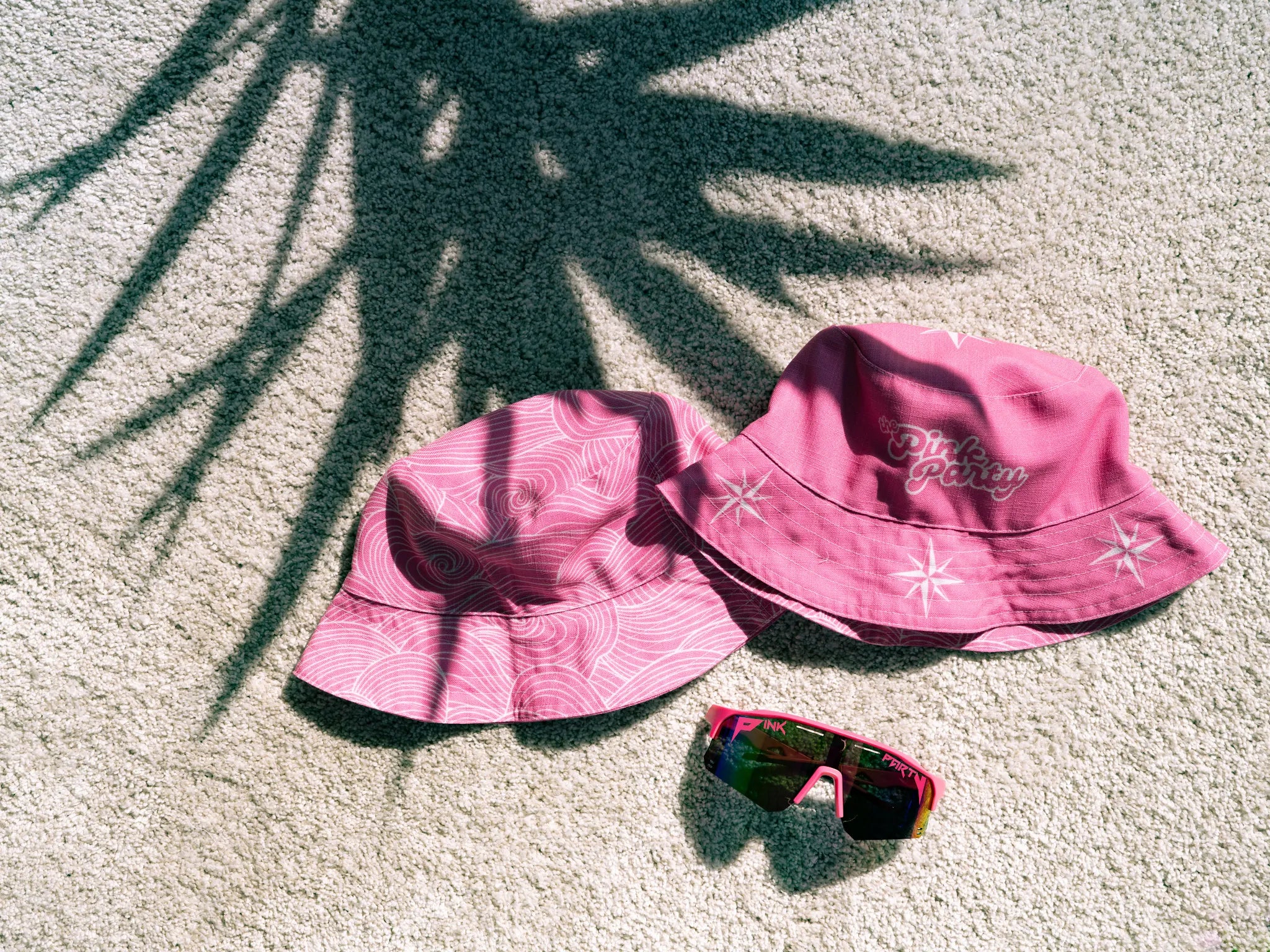 Two pink bucket hats and a pair of sunglasses on a textured surface with palm leaf shadows.